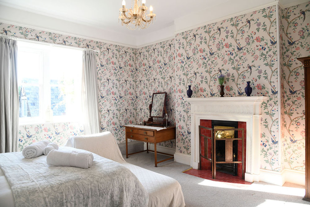 An interior shot of the King bedroom at Lorton House with flowery wallpaper, a feature fireplace and dressing table. The bed is dressed with towels and a chaise lounge is at the end of the bed.