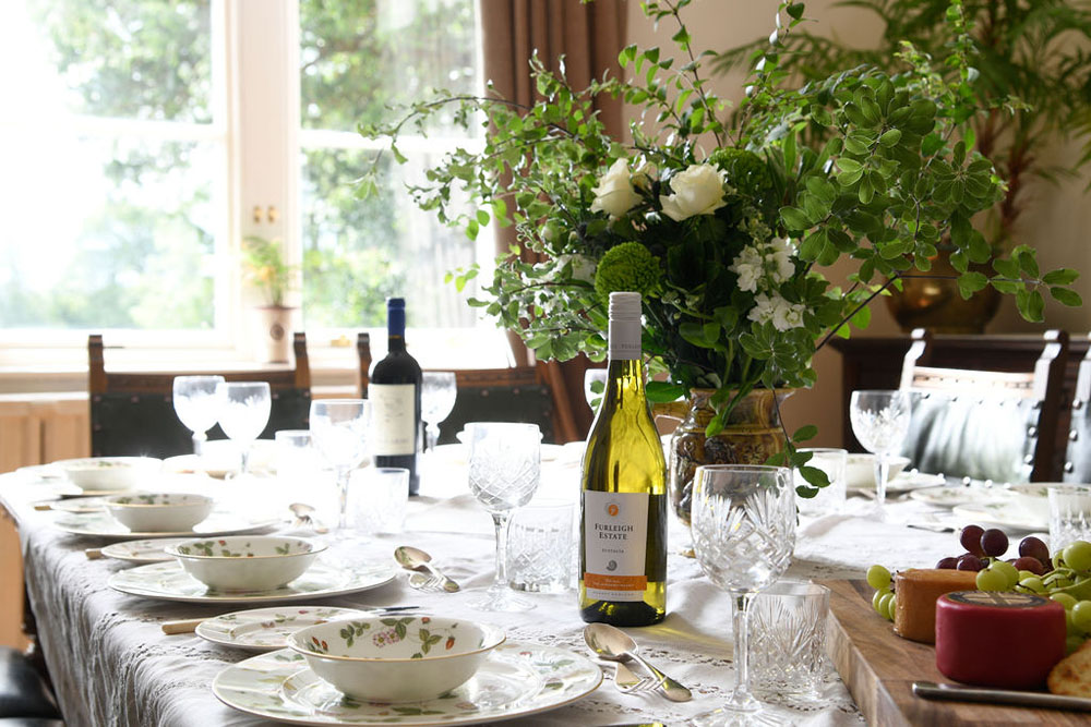 The dining room at Lorton House. The large wooden table is set with bowls, plates and engraved glasses with a bottle of white wine from Furleigh Estate and a large bunch of flowers in the centre of the table.