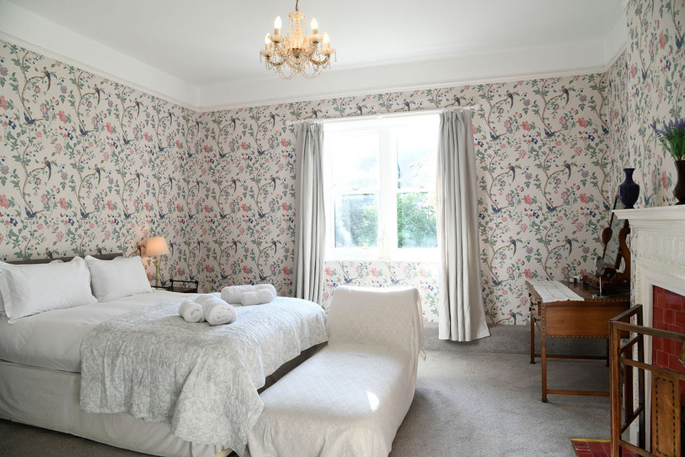 A bedroom at Lorton House featuring floral wallpaper, a large bed, chase lounge, desk and fireplace.